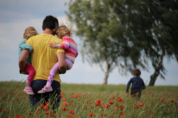 Ein Mann tr&auml;gt zwei kleine Kinder auf dem Arm, ein drittes Kind l&auml;uft vor  ihm durch eine Mohnblumenwiese.